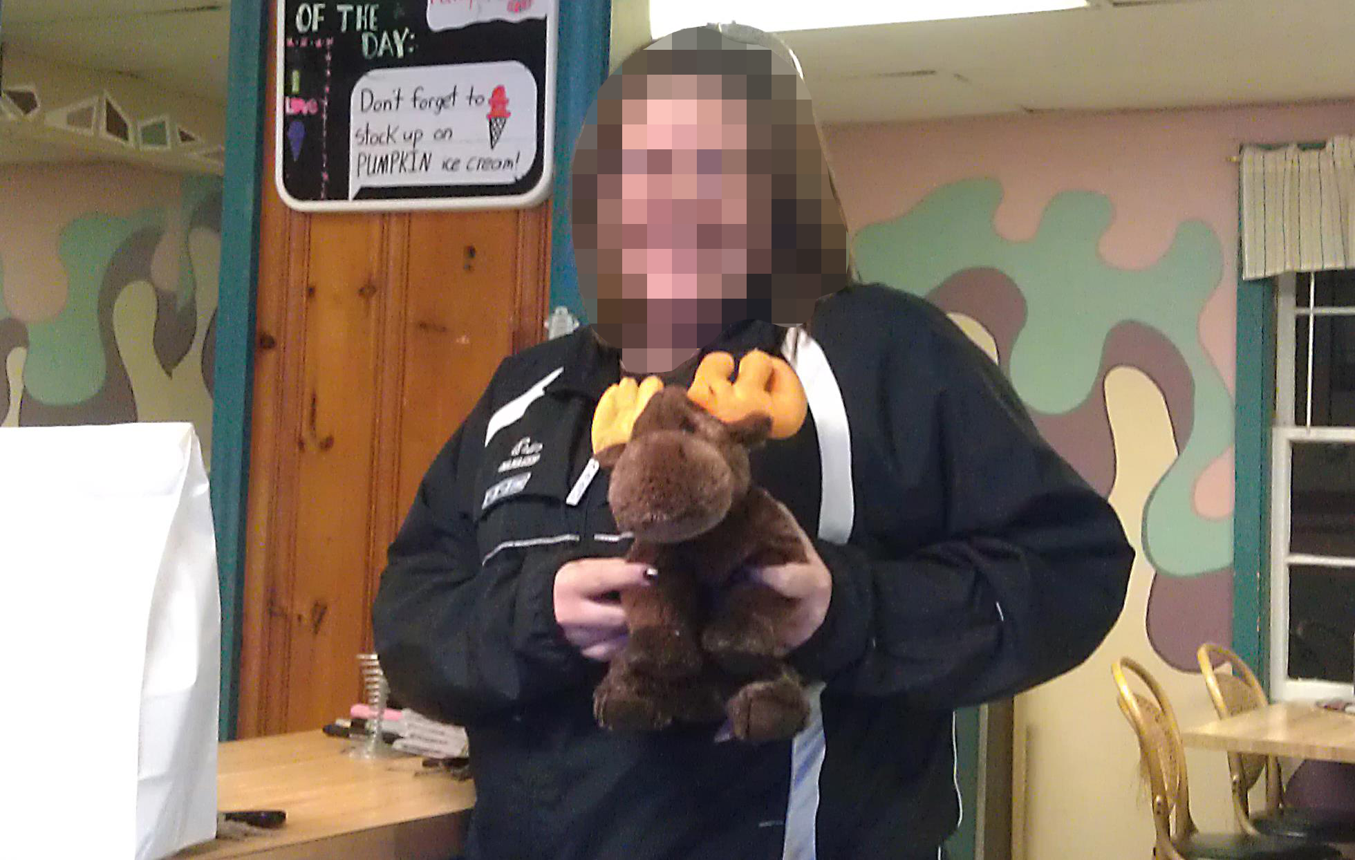 A light-skinned woman with her face blurred is holding a stuffed moose. The background has walls that are cream, purple, green, and pink colored splotches.