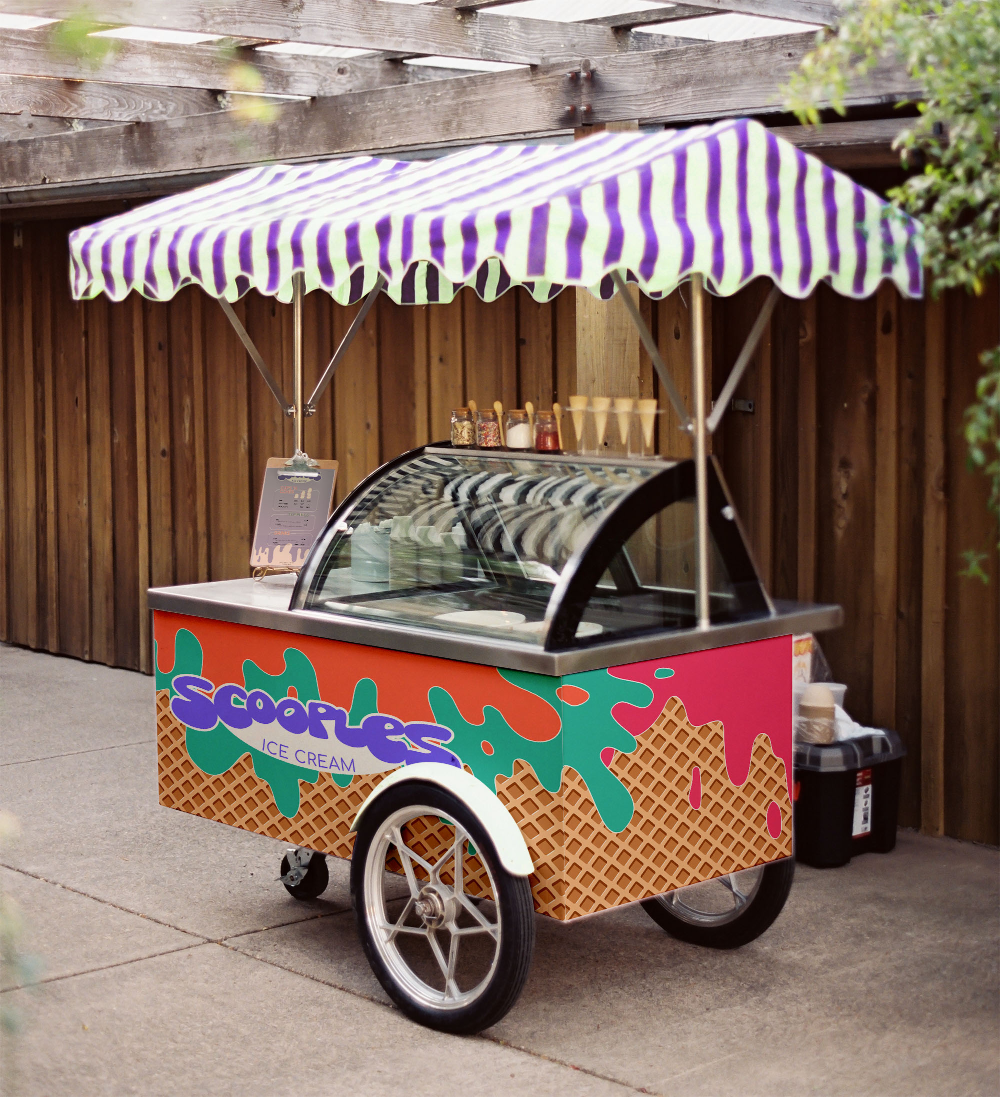 A small ice cream cart with a waffle cone pattern, pink, green, and orange splotches. The canopy is white and purple striped.